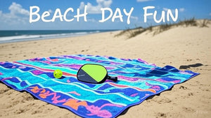 A vibrant Hatteras beach scene with a pickleball paddle and ball on a colorful towel, sandy dunes, and gentle waves under a bright blue sky, capturing the laid-back vacation spirit.